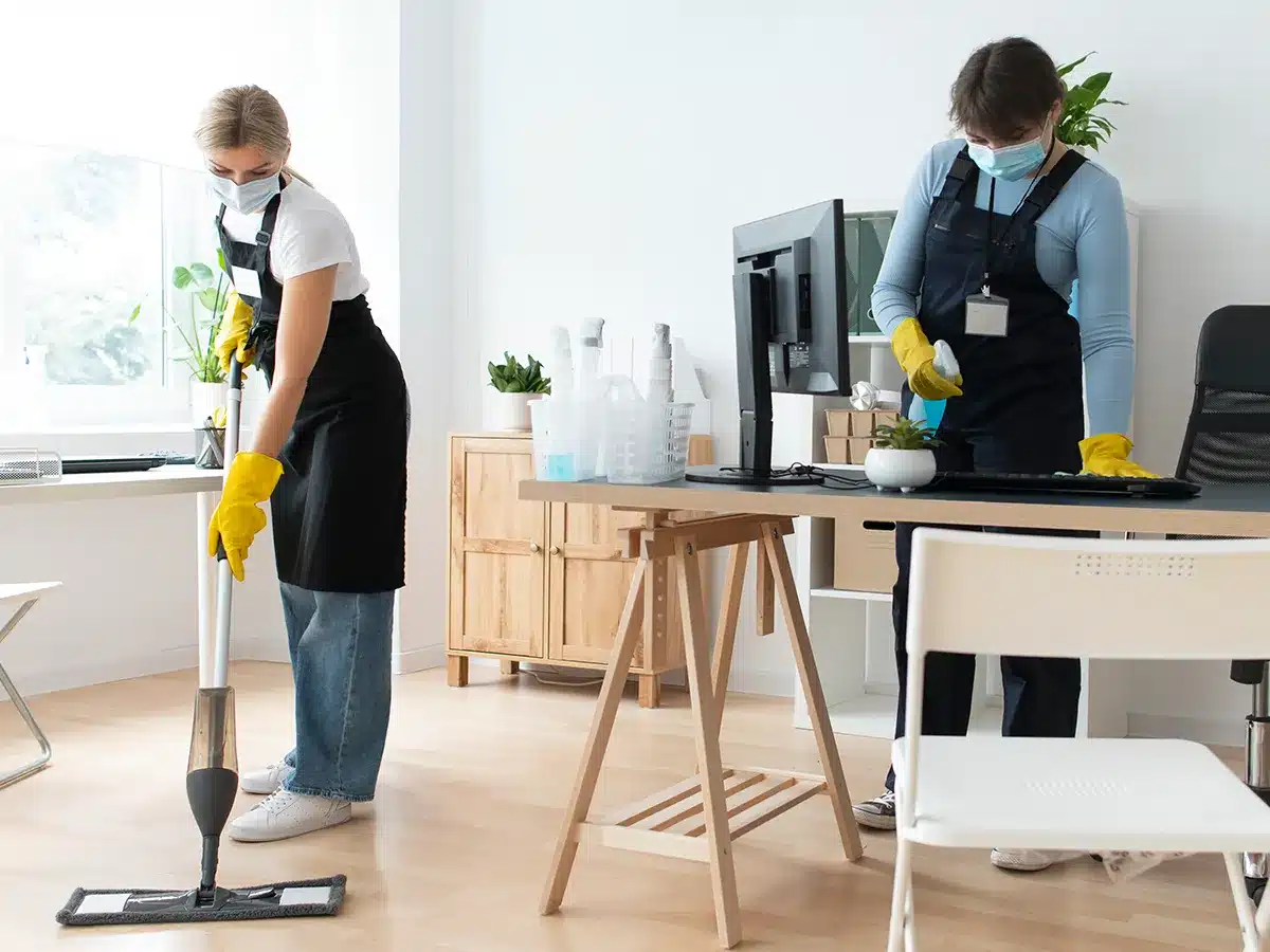 Reinigungskräfte während der Büroreinigung. Eine Mitarbeiterin wischt den Boden und eine andere Reinigungskraft macht den Tisch sauber.